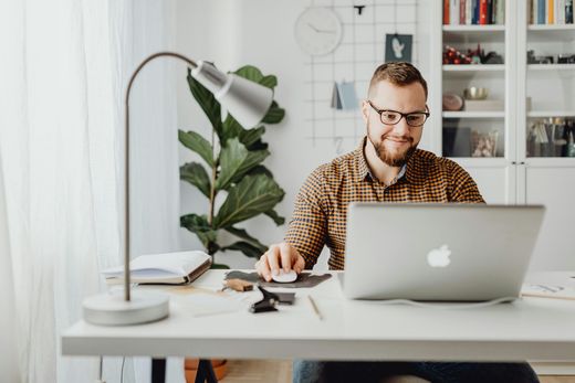 Un homme avec des lunettes utilisant un ordinateur portable à un bureau, souriant, avec une plante et une lampe.