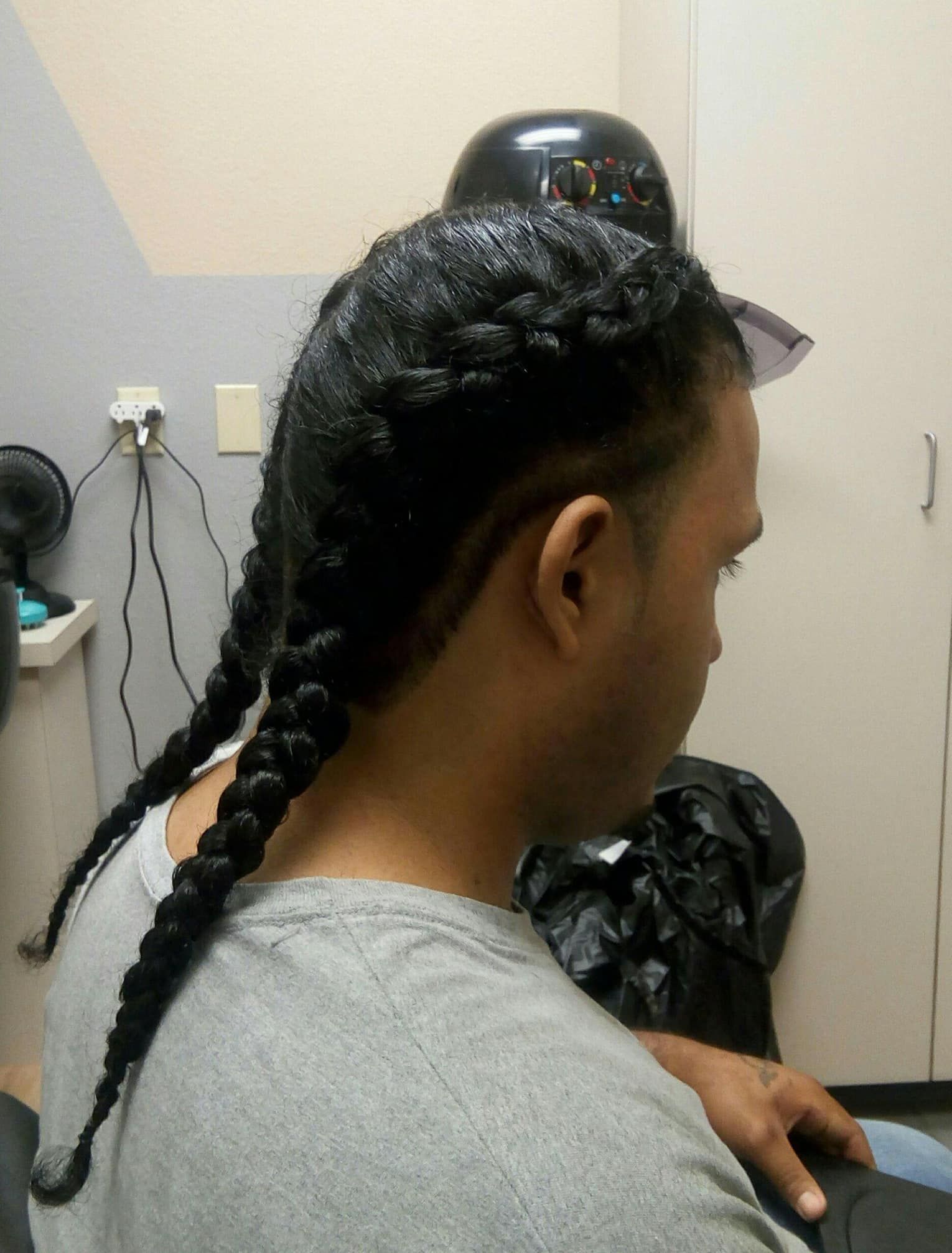 Man with dark hair in two French braids, wearing a gray shirt, in an interior setting.