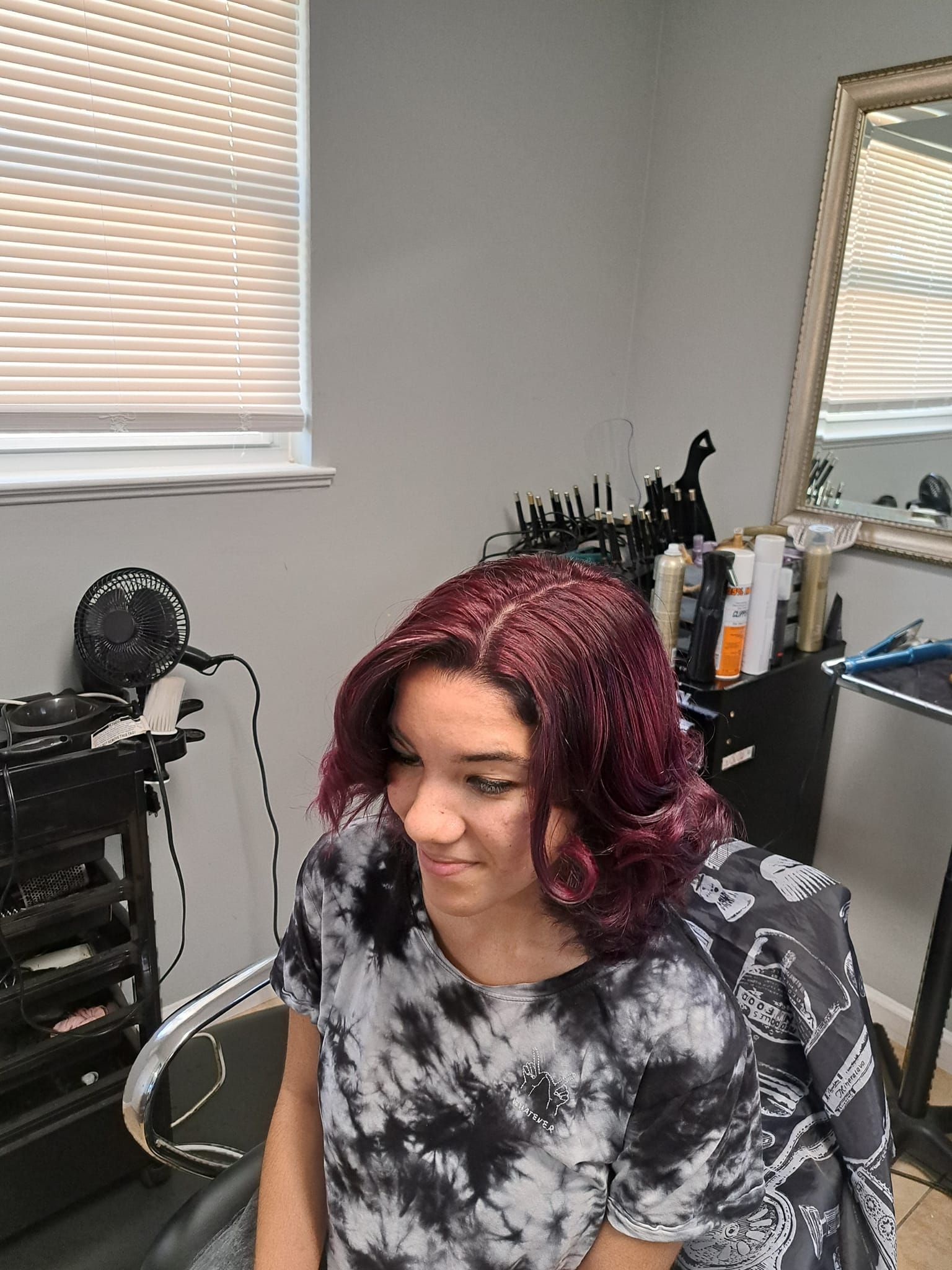 Woman with dark red wavy hair in a salon chair, wearing a tie-dye shirt.