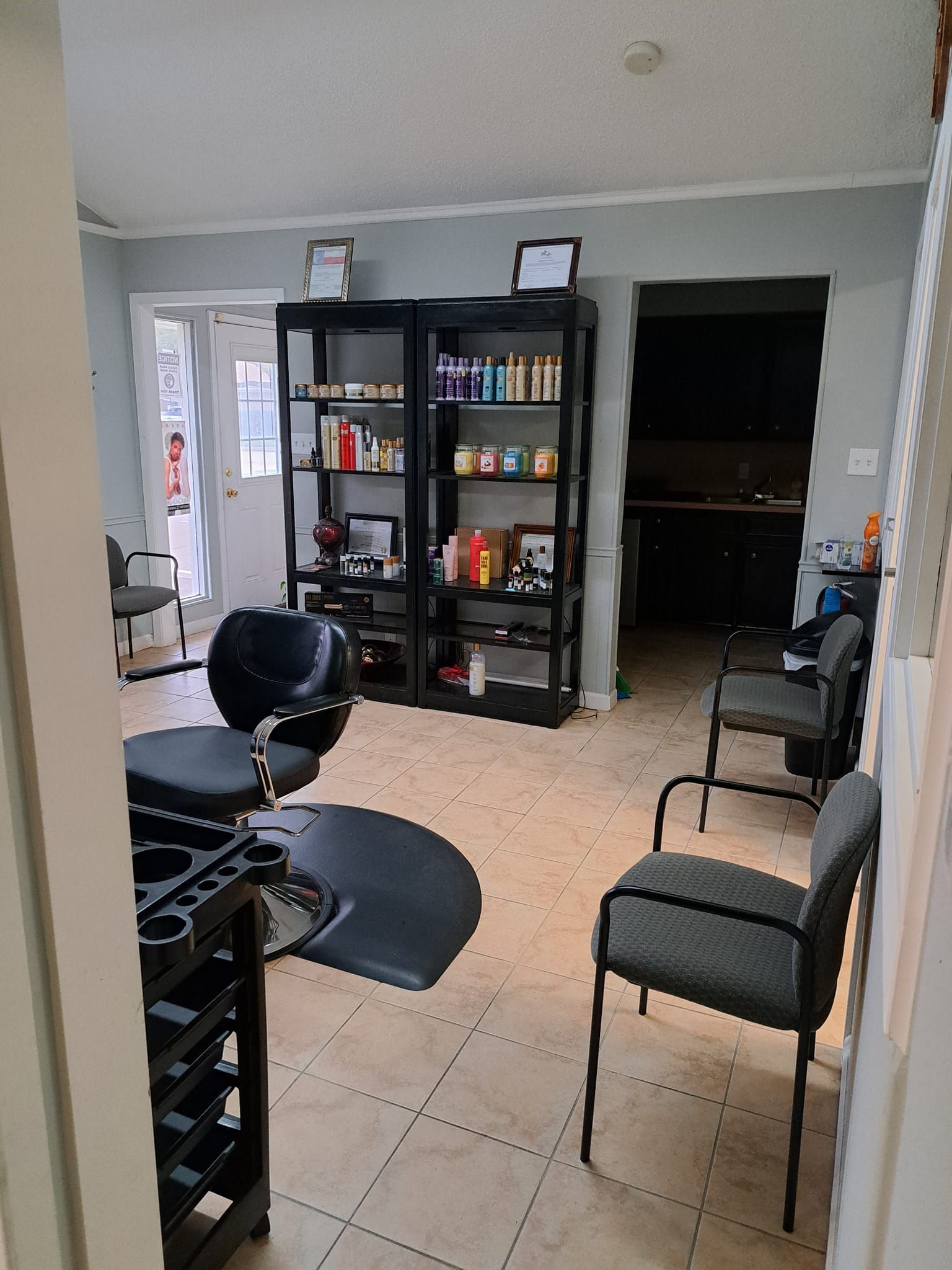 Hair salon interior: black styling chair, product shelves, waiting area with chairs, doorway to kitchen.