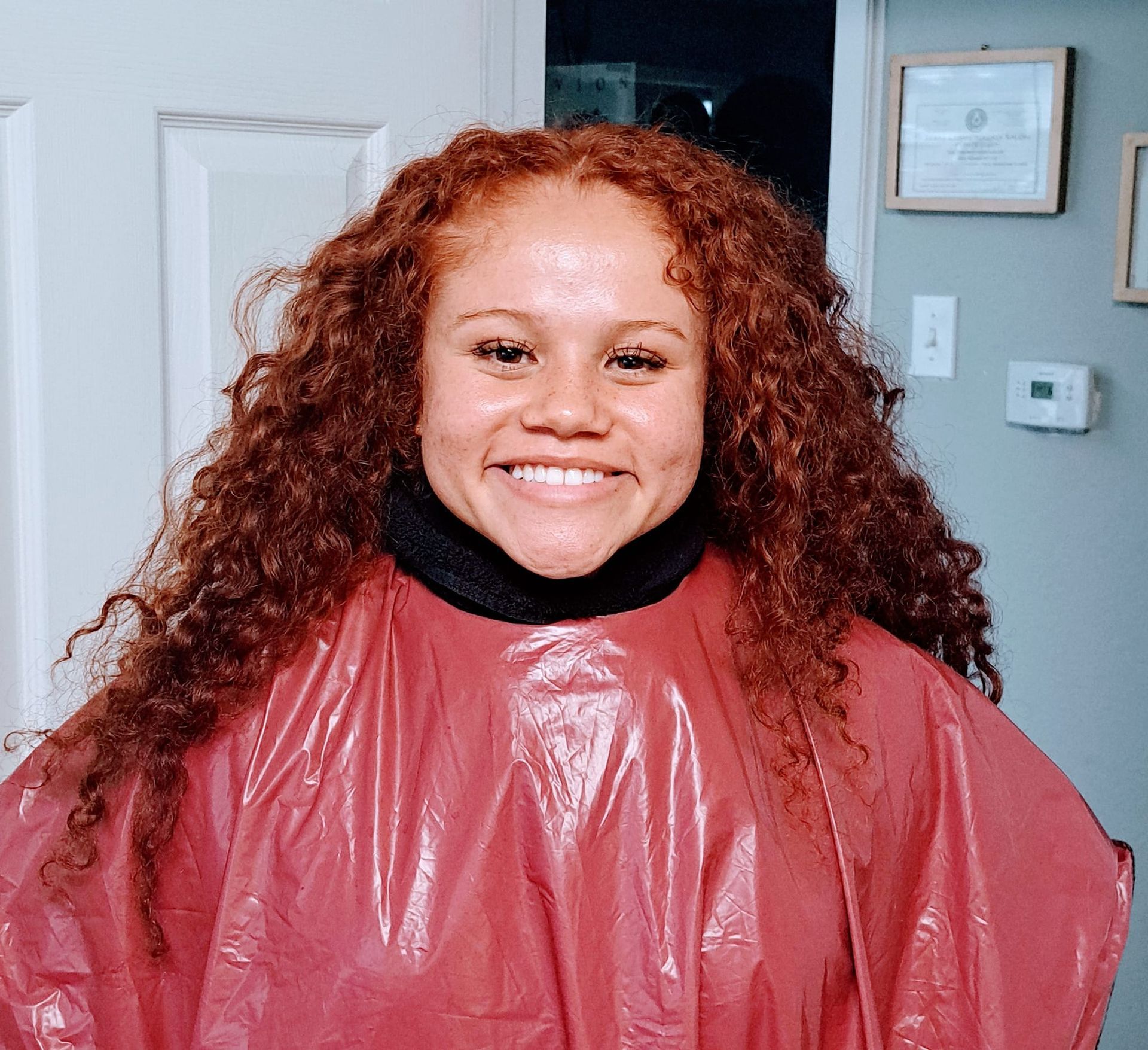 Woman with long, curly auburn hair, smiling, wearing a black top and red salon cape.