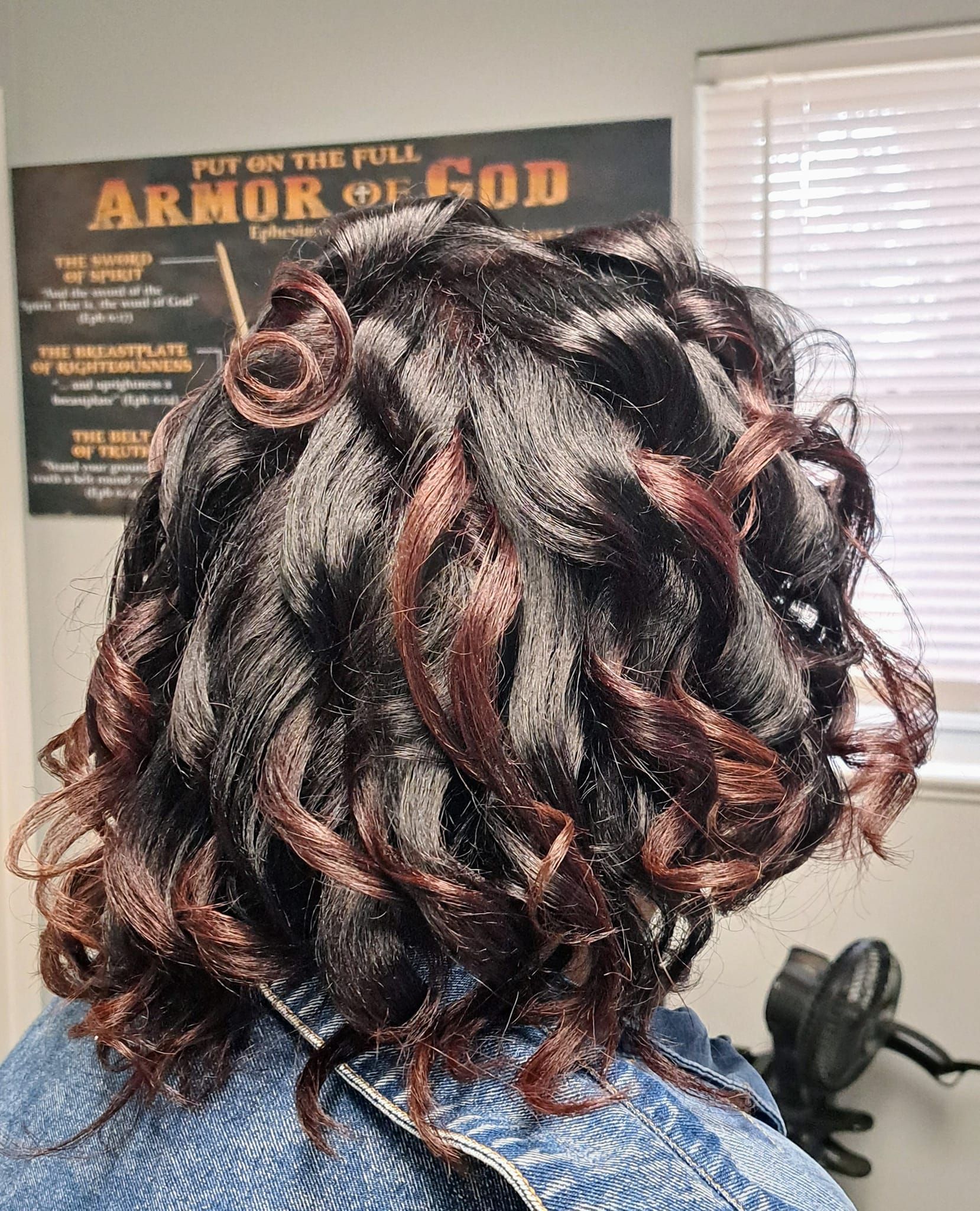 Woman's dark hair styled with curled sections. Highlights of reddish-brown. Blue shirt, wall with blind.
