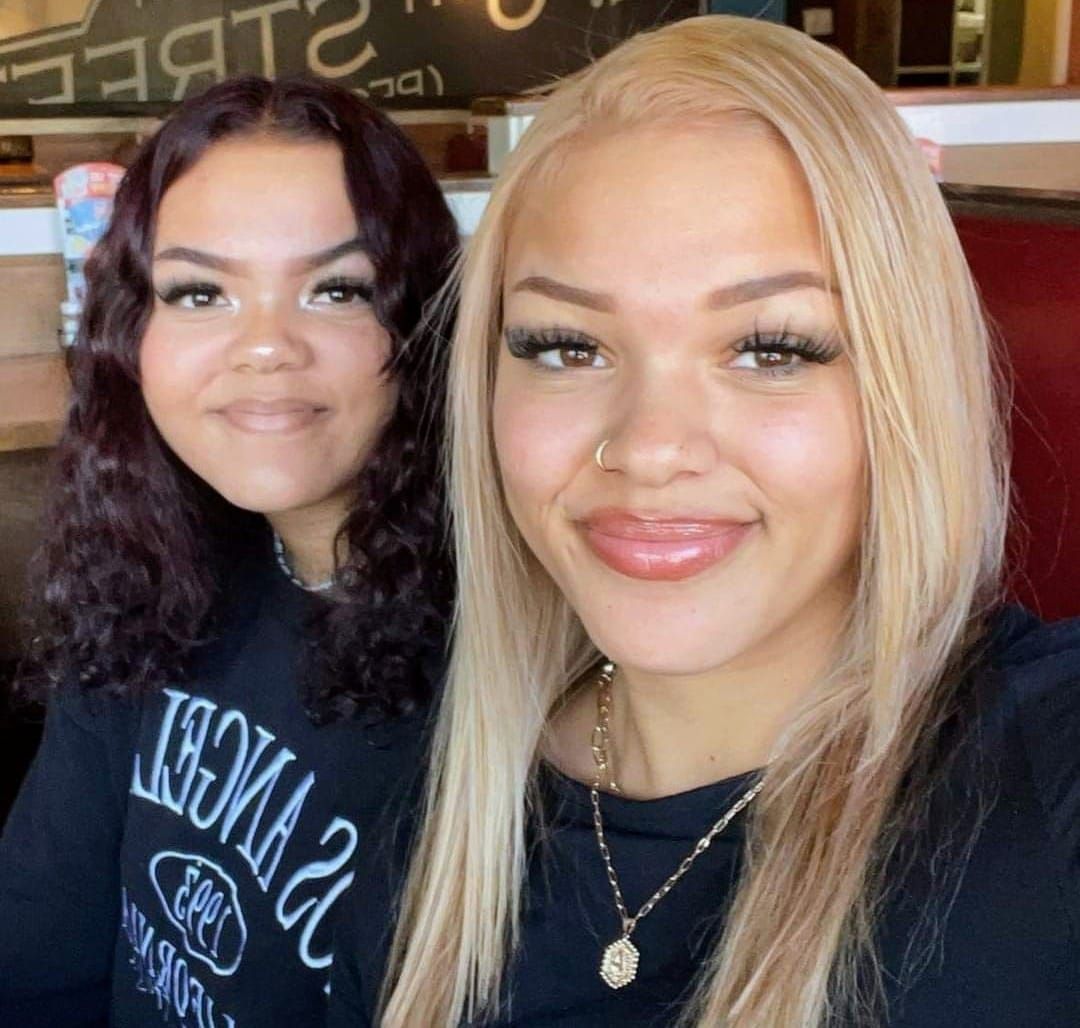 Two women smiling; one with blonde hair, the other with dark hair. Indoors, seated.