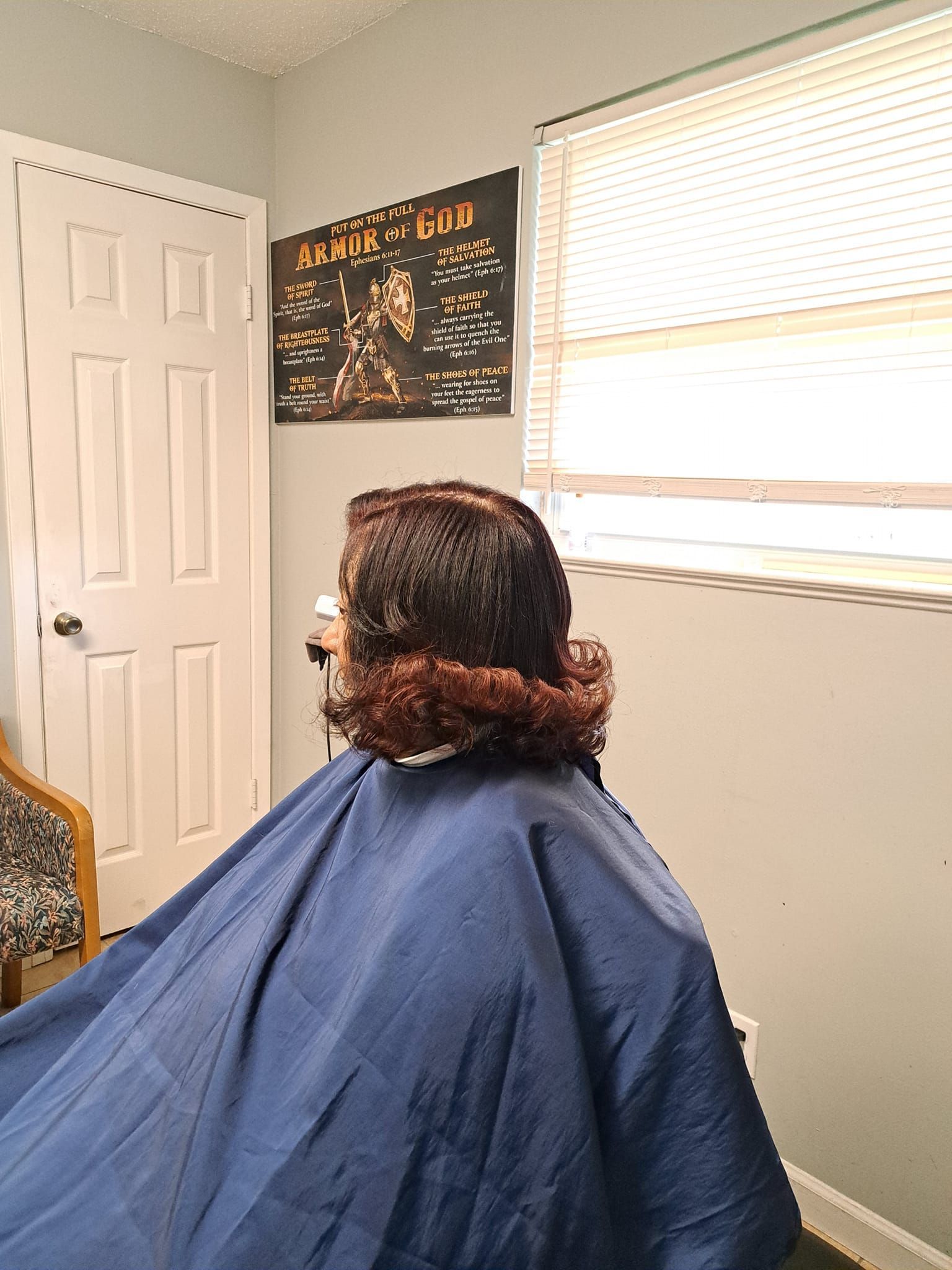 Person in a blue cape, getting hair styled in a room with a closed white door and a poster on the wall.