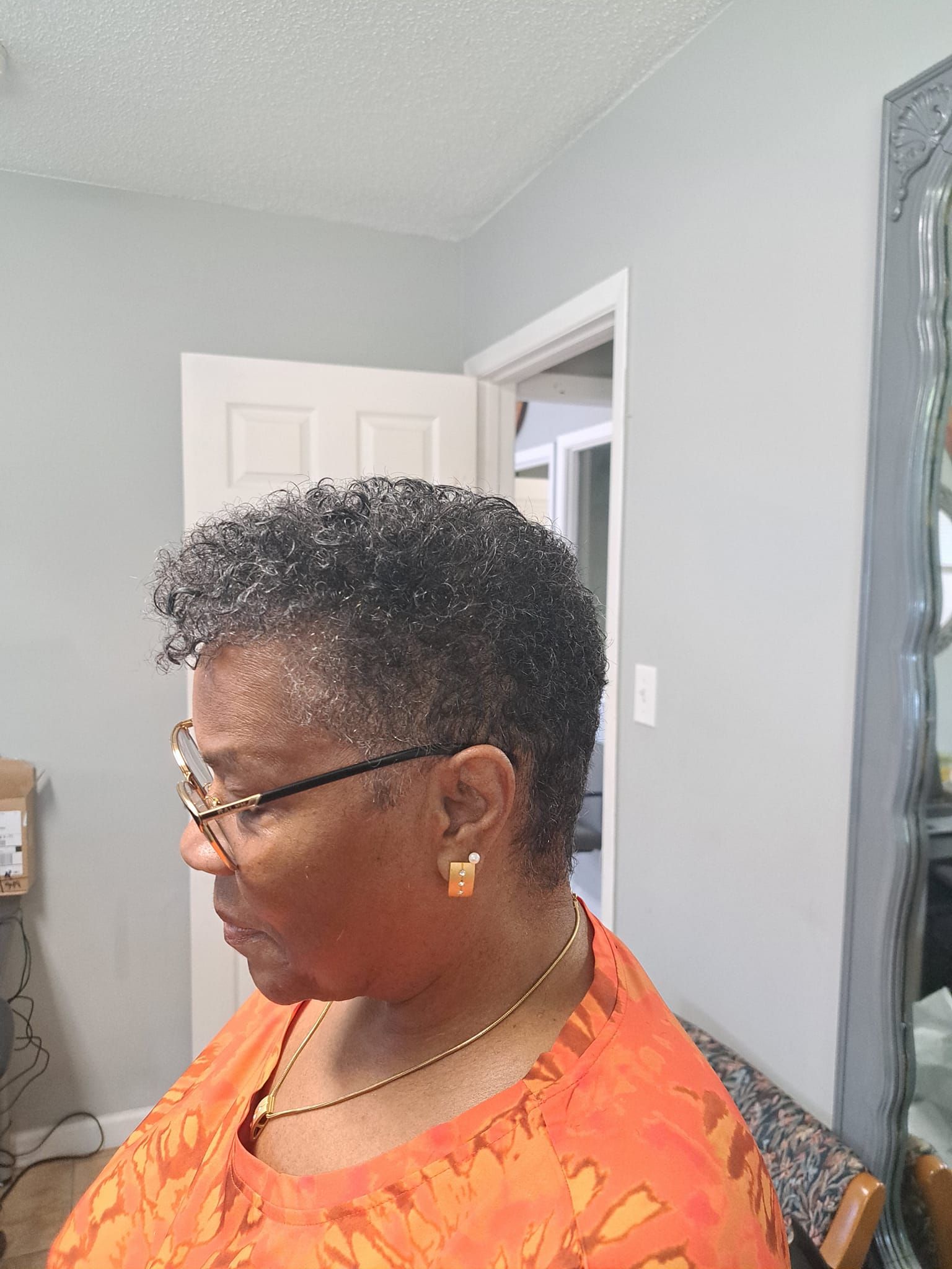 Woman with glasses and short, curly dark hair wearing an orange patterned shirt, sitting indoors.