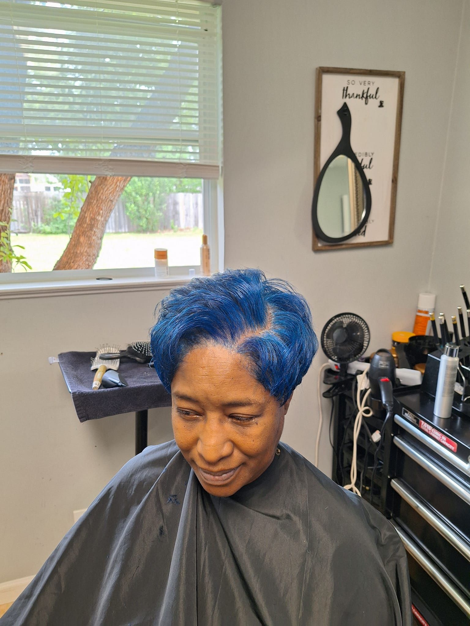 Woman with short, blue hair in a salon chair, smiling. Near a window and a mirror decoration.