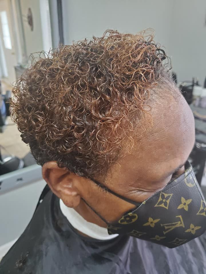 Person with short, curly, brown hair, wearing a patterned face mask, sitting in a salon chair.
