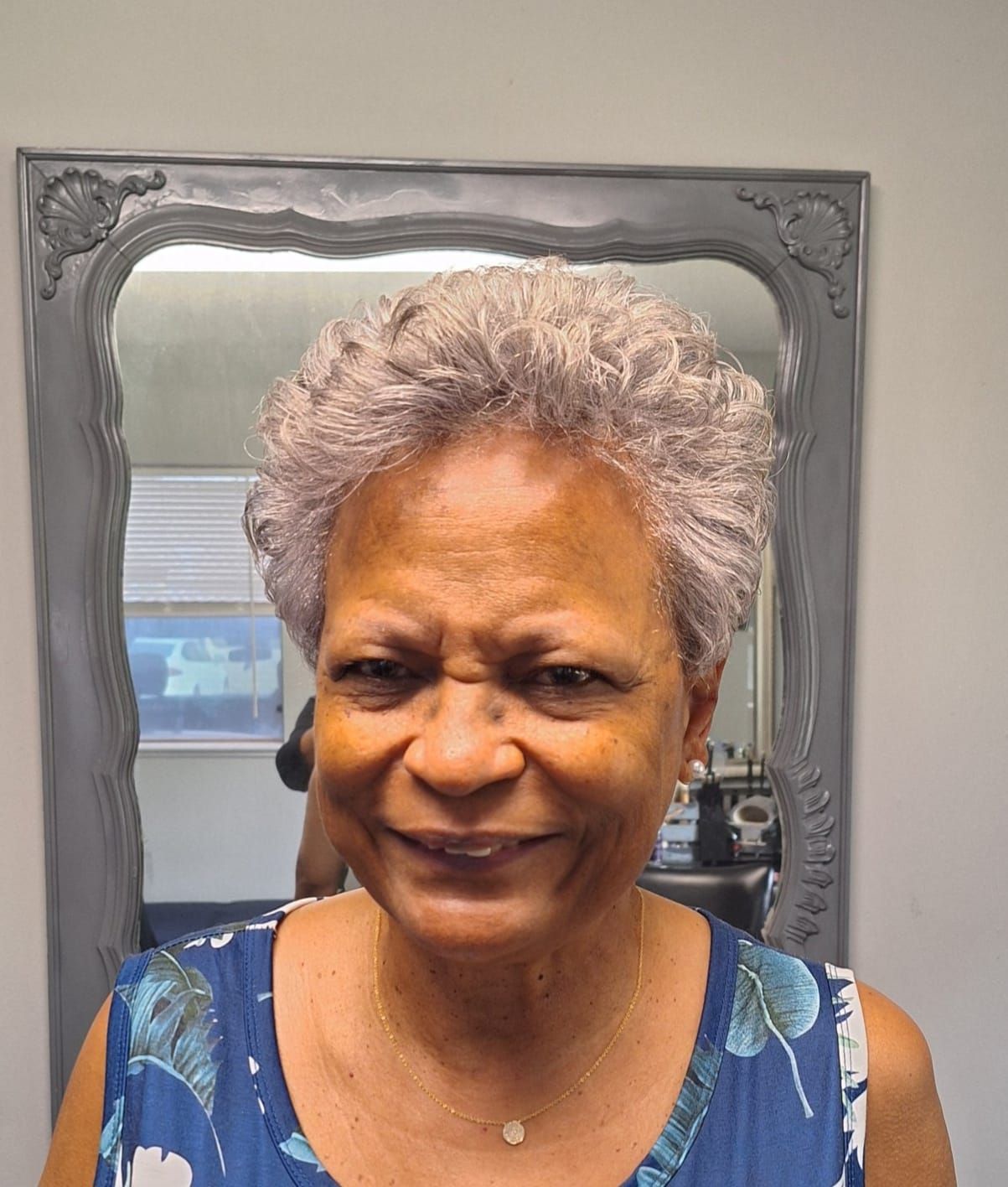 Woman with short, gray curly hair smiles in front of a mirror, wearing a blue floral top.