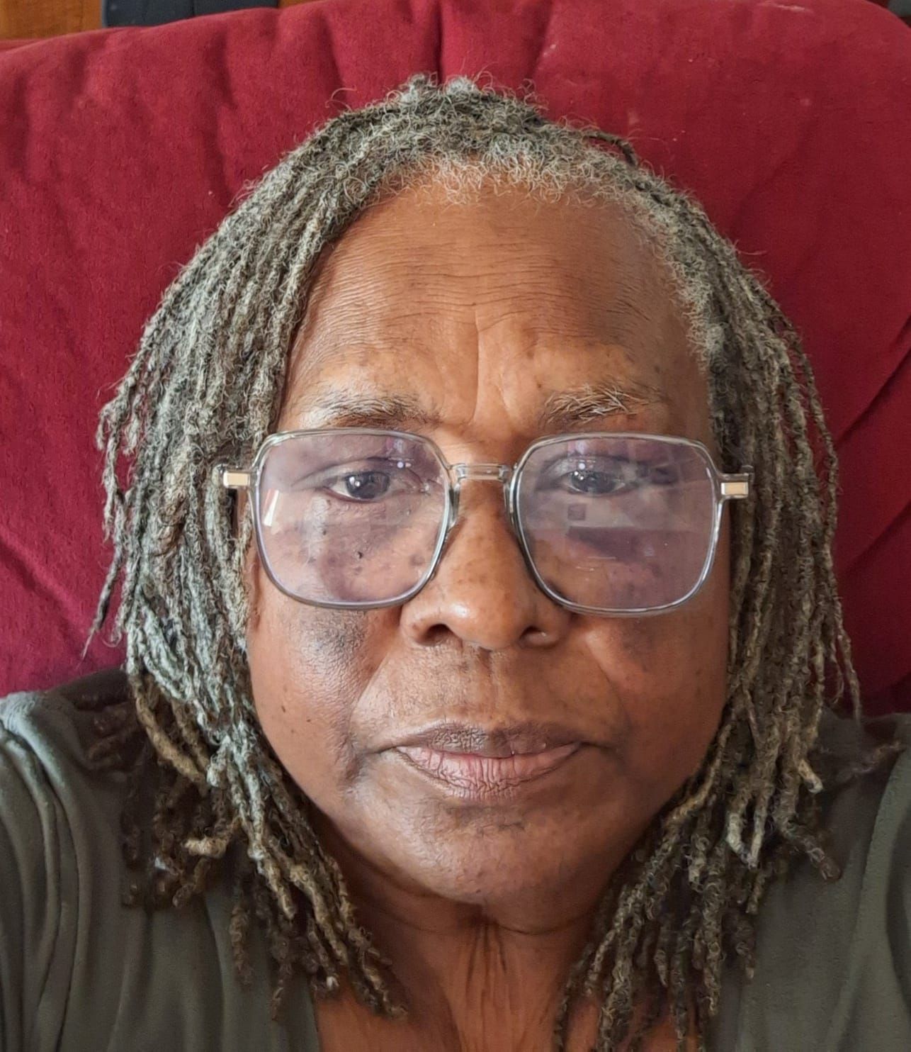 Person with dreadlocks and glasses, looking at the camera. Wearing a green shirt and sitting.