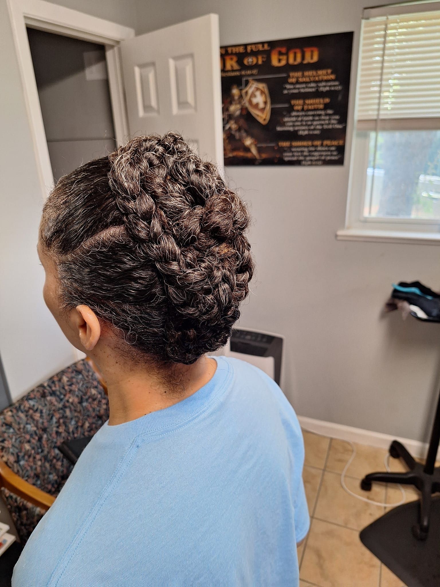 Woman with dark, textured hair styled up, wearing a light blue shirt. Indoor setting with a poster and window.