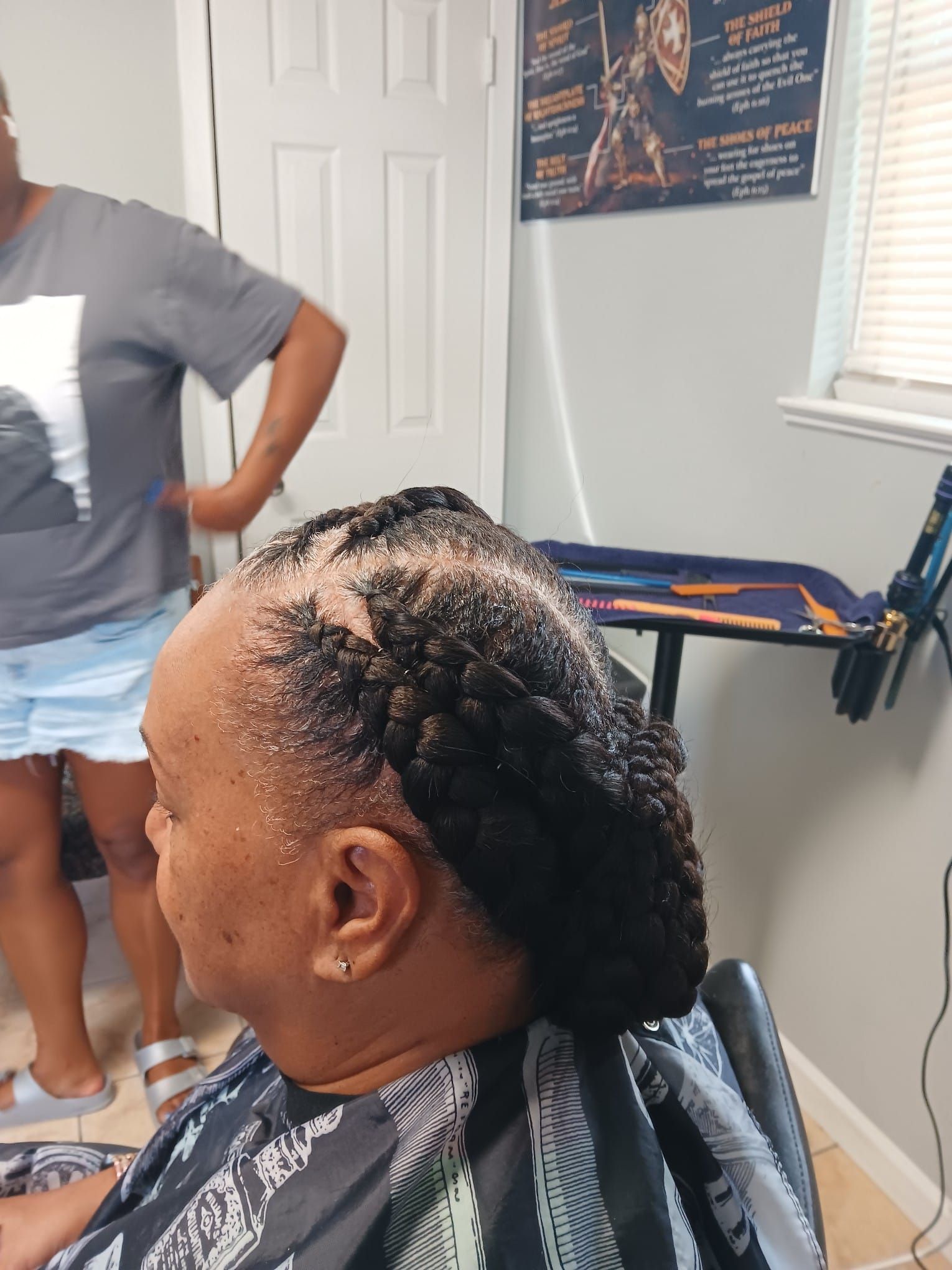 Woman with braided hairstyle in a salon chair; another woman in a gray shirt stands behind.