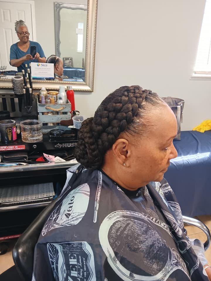 Woman with braided updo in a salon. Another person is reflected in the mirror, taking a photo.