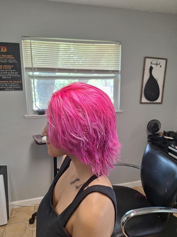 Woman with bright pink hair in a salon; she sits in a chair near a window.