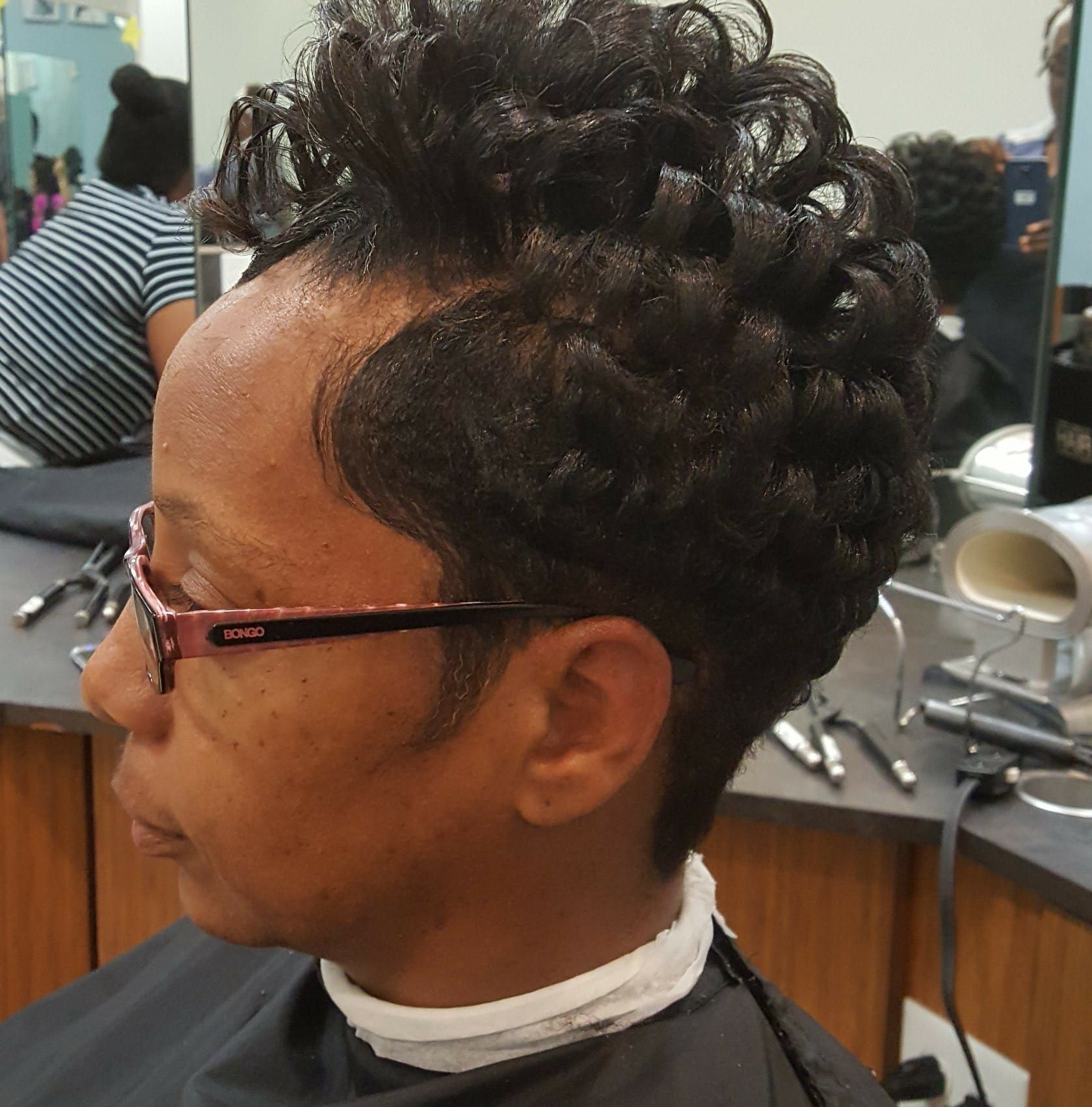 Woman with short, dark, curly hair in a salon, wearing glasses and a salon cape.