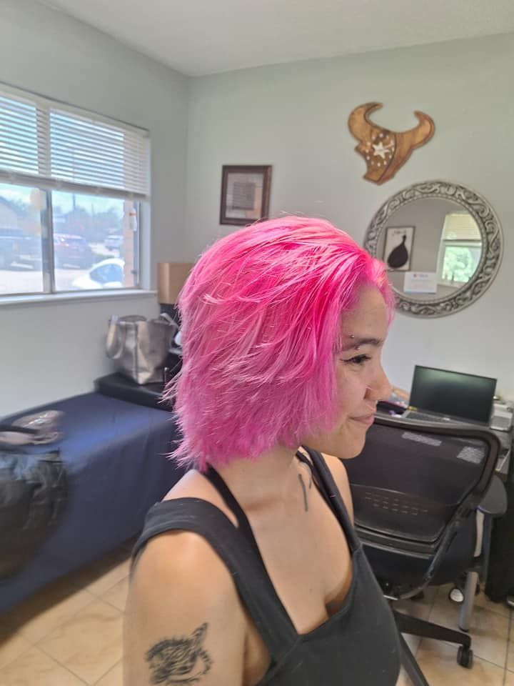 Woman with bright pink hair in a salon. Wearing a black tank top, facing right, smiling slightly.