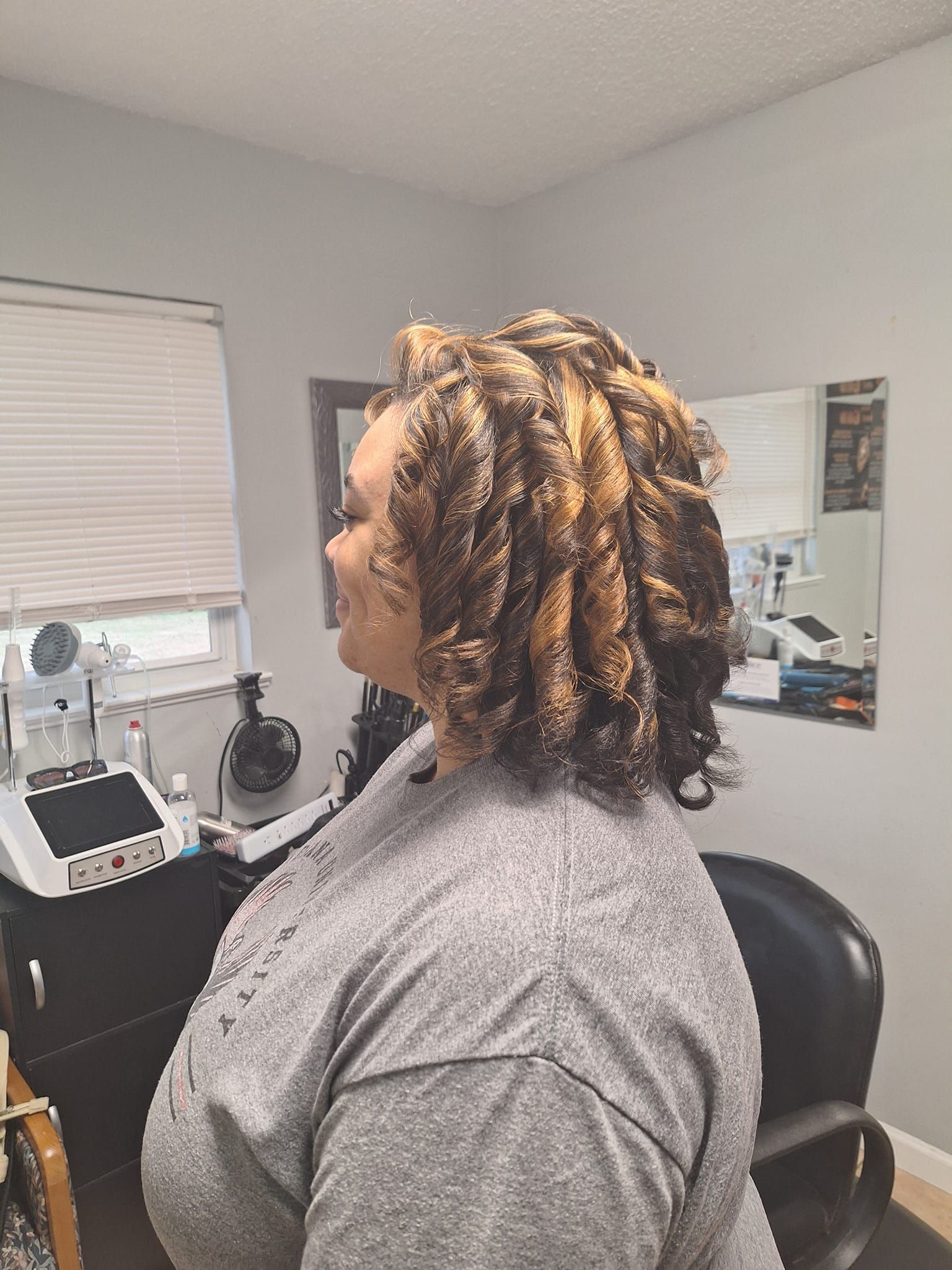 Woman with shoulder-length, curled hair, wearing a gray shirt, smiles in a salon, a mirror is behind her.