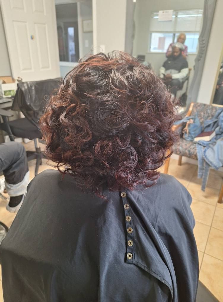 Person with dark, curly, auburn-tinged hair in a salon chair, wearing a black cape.