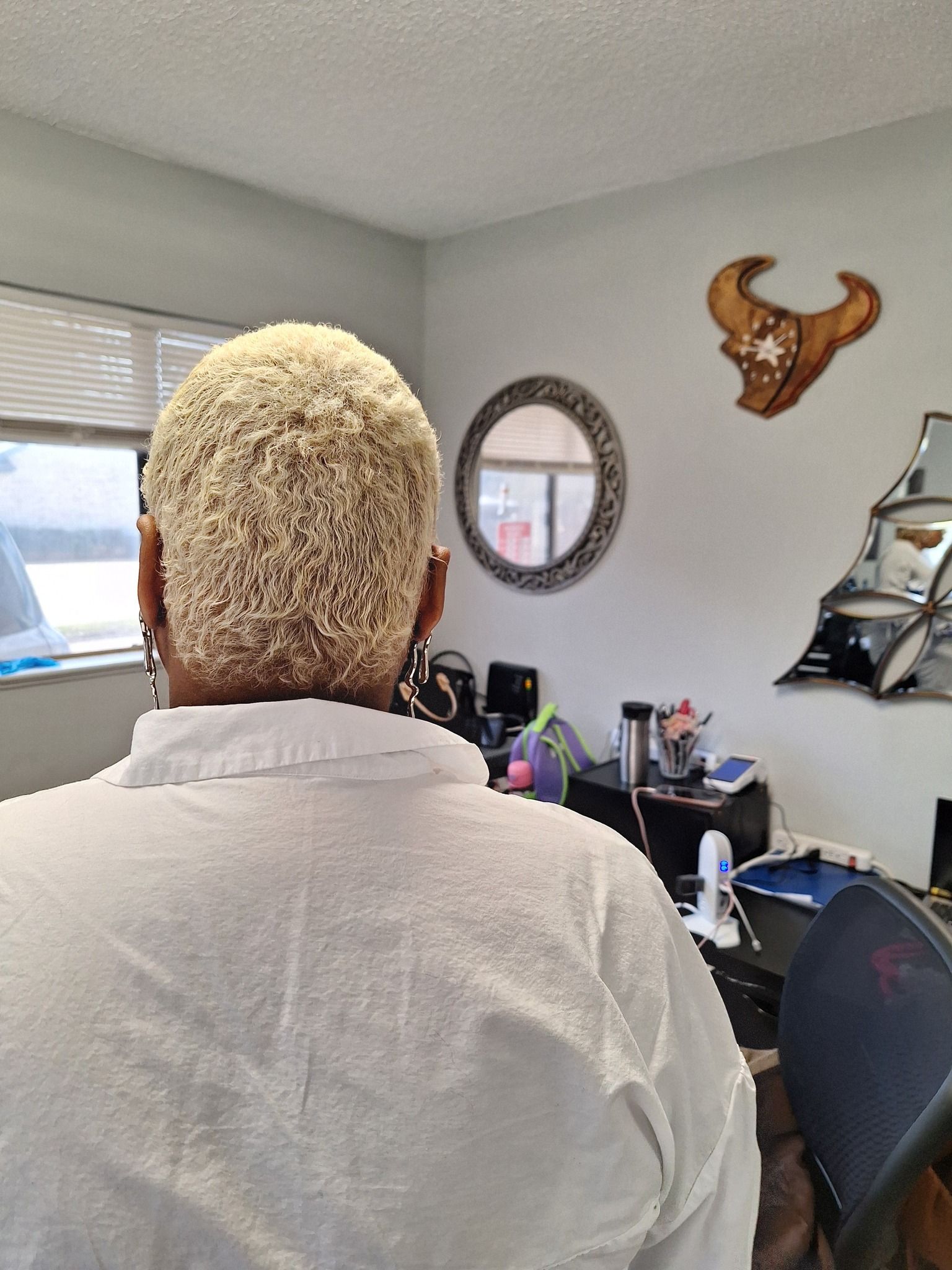 Woman with blonde, textured hair, wearing a white shirt, in a room with a mirror and decor.
