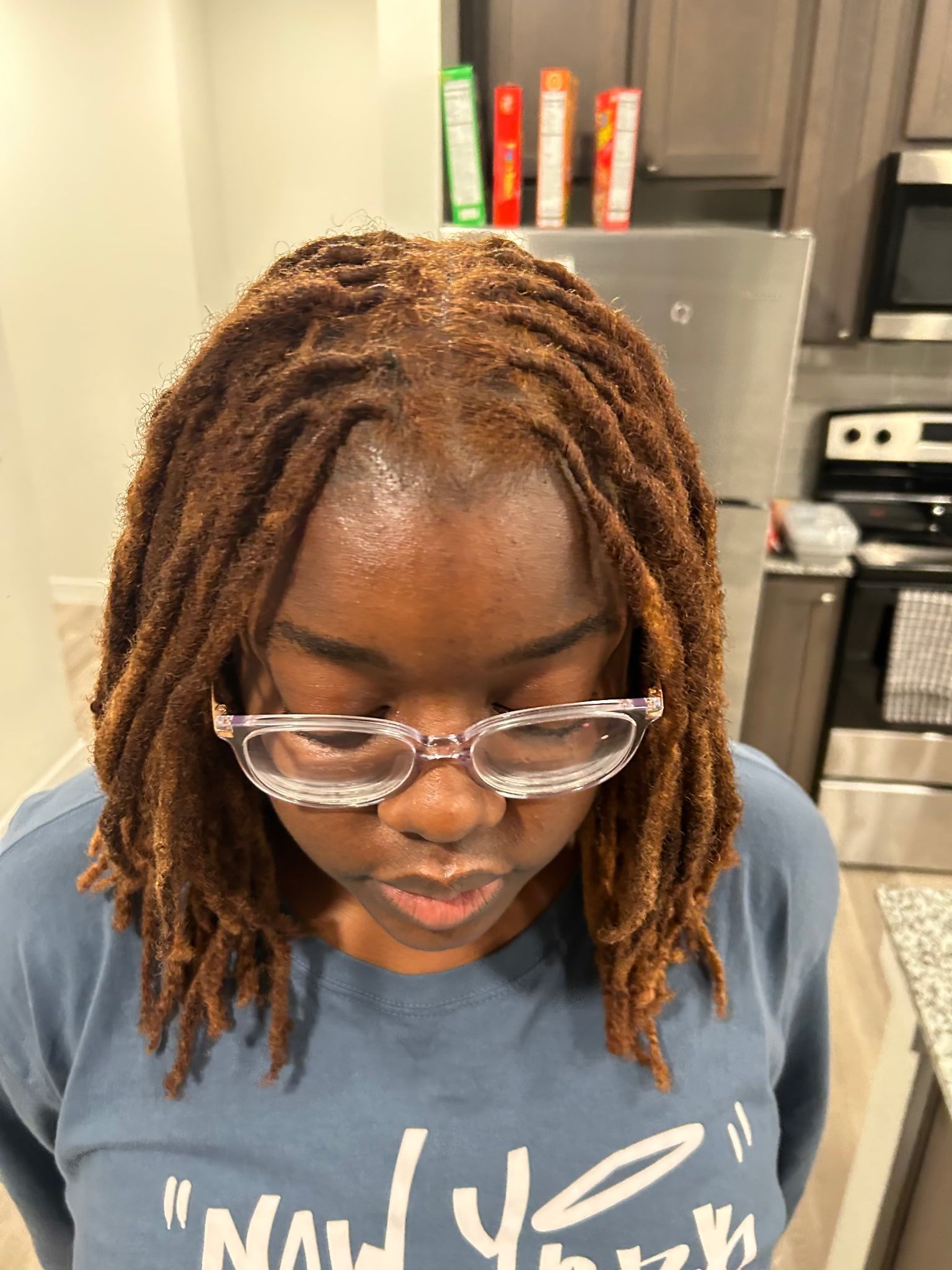 Person with brown dreadlocks and glasses looking down in a kitchen setting.