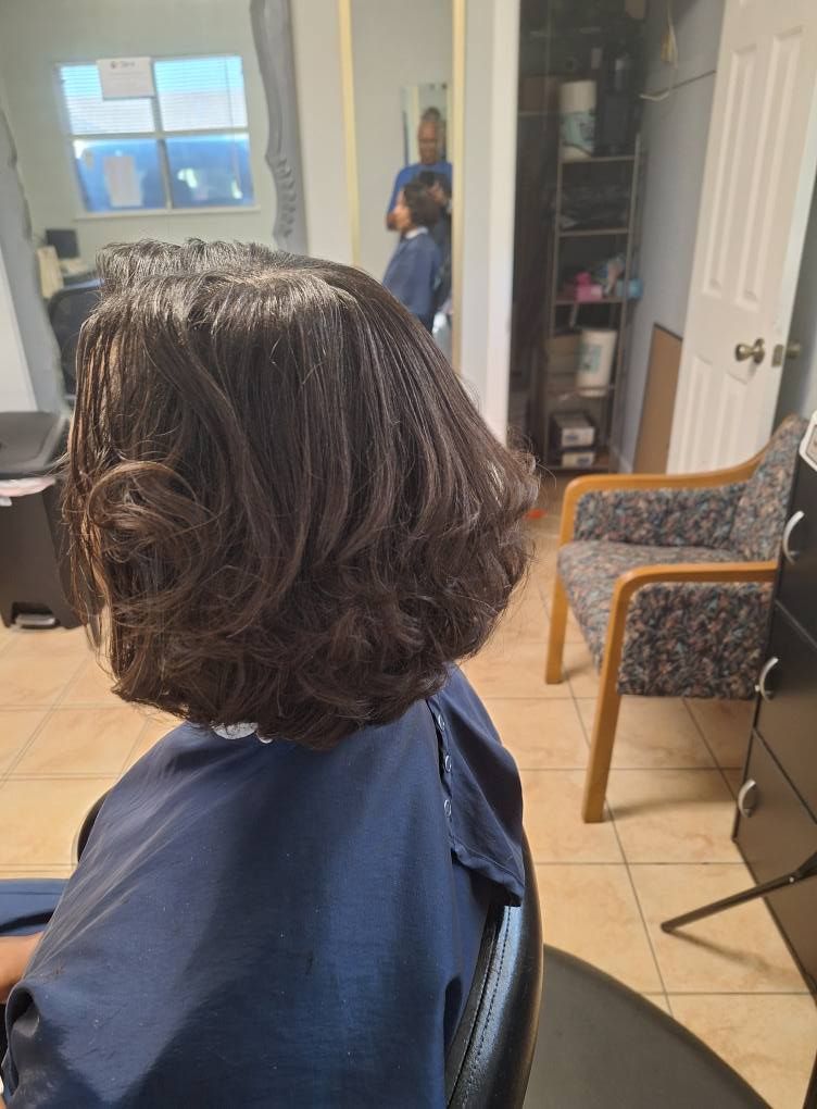 Person in a salon chair with layered dark hair; blue salon cape. Interior shot with mirror.