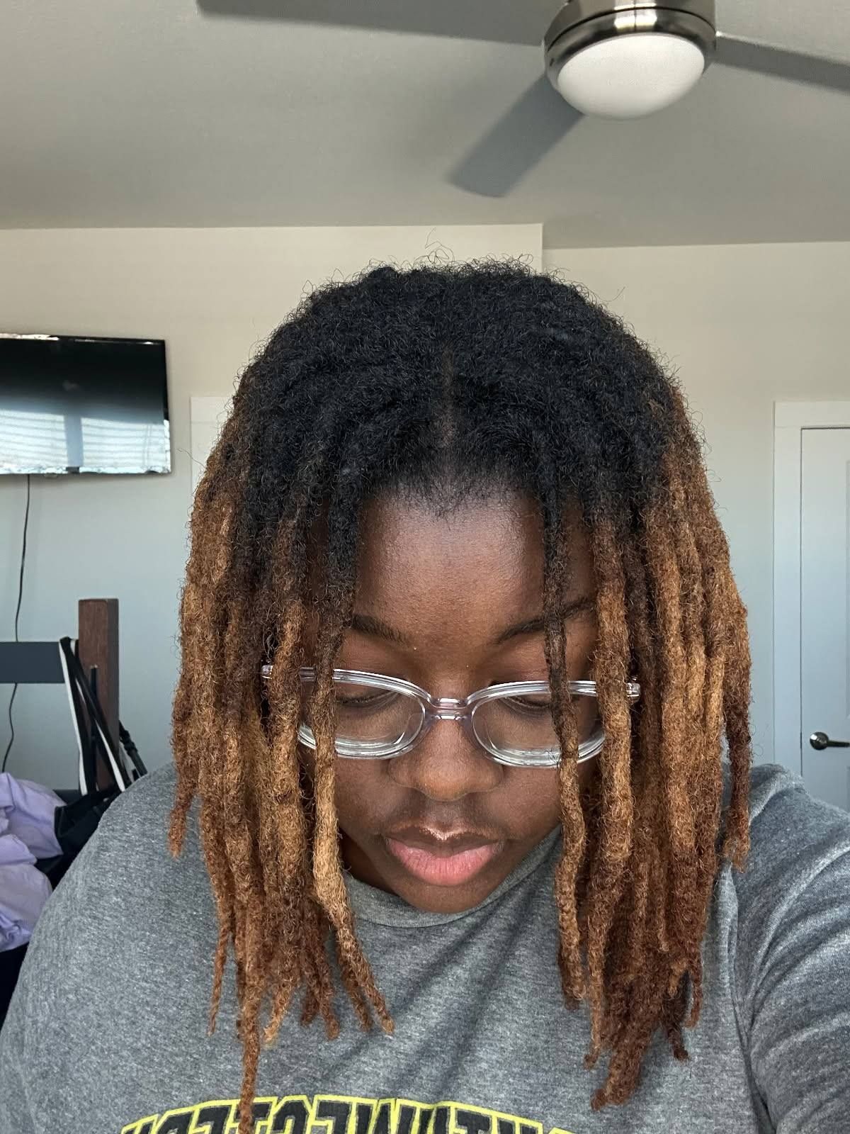 Person with locs, ombre blonde ends, wearing glasses. Indoors, looking down.
