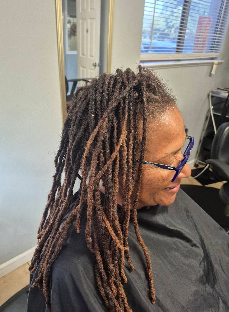 Woman with brown dreadlocks and blue-framed glasses, smiling, wearing a black cape, indoors.