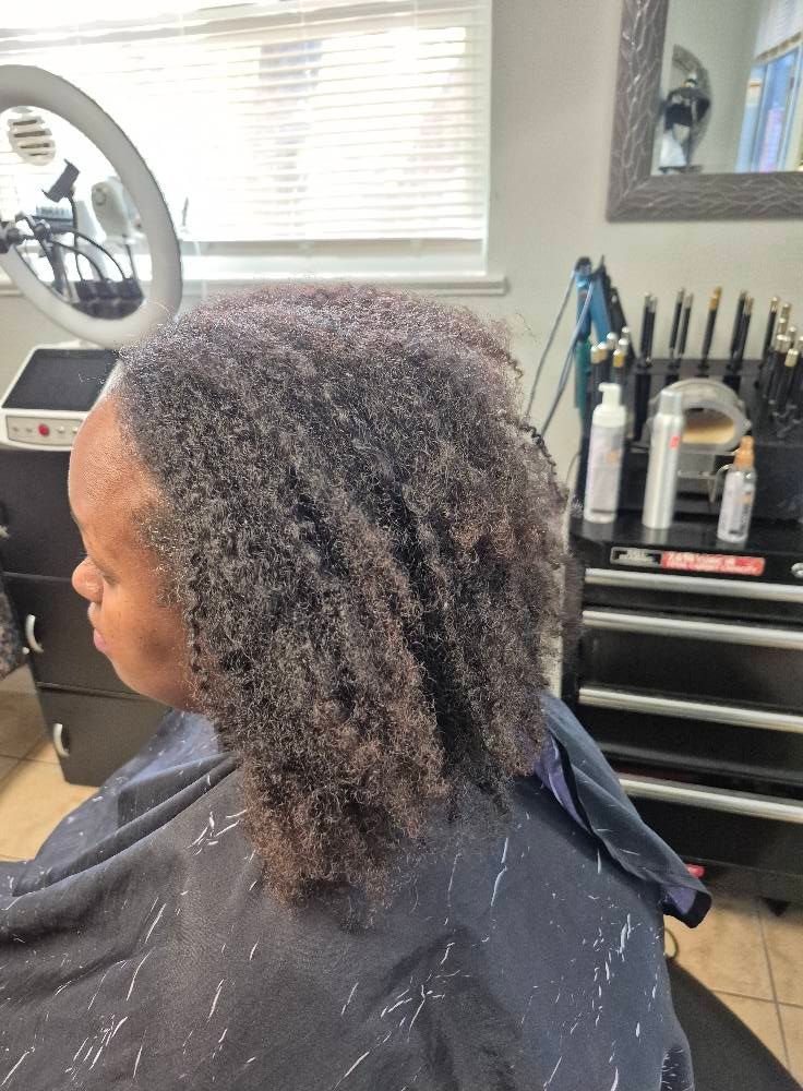 Person with textured hair at a salon, wearing a cape. Hair is partially styled, with styling tools in the background.