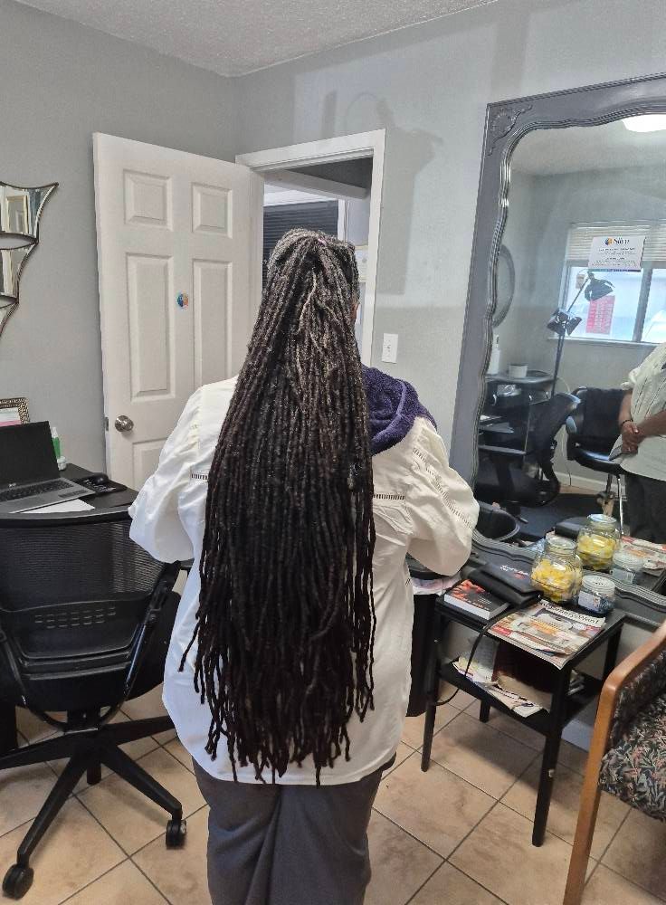 Person with long dreadlocks standing in a room with a mirror and furniture.