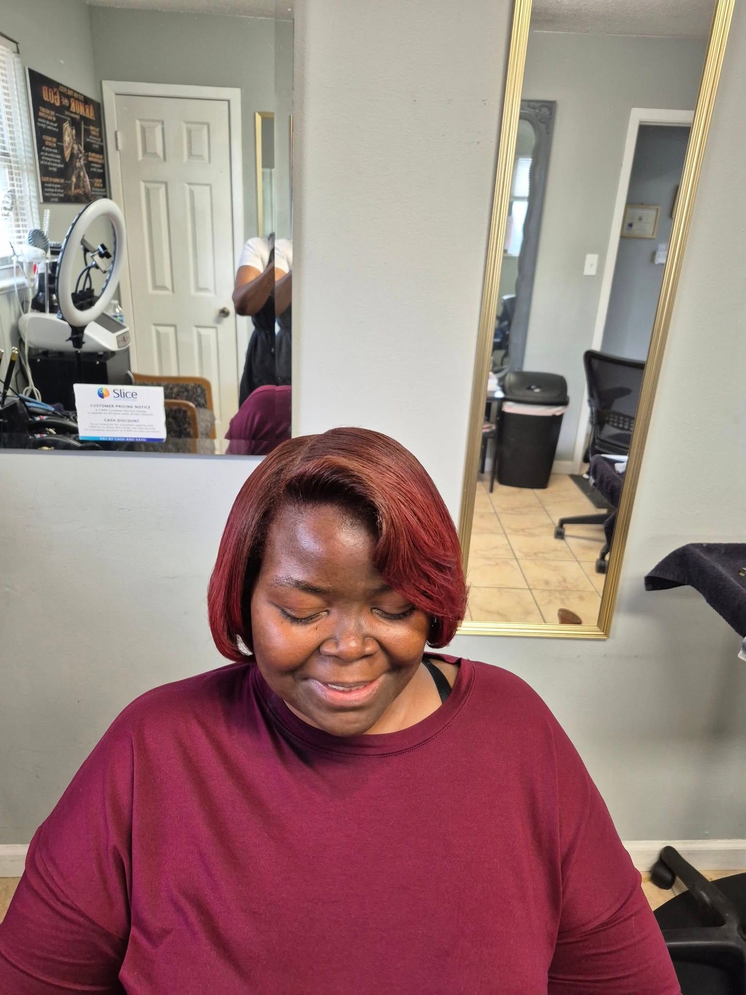 Woman with reddish-brown hair smiles in a salon, wearing a burgundy sweater. She is near a mirror.