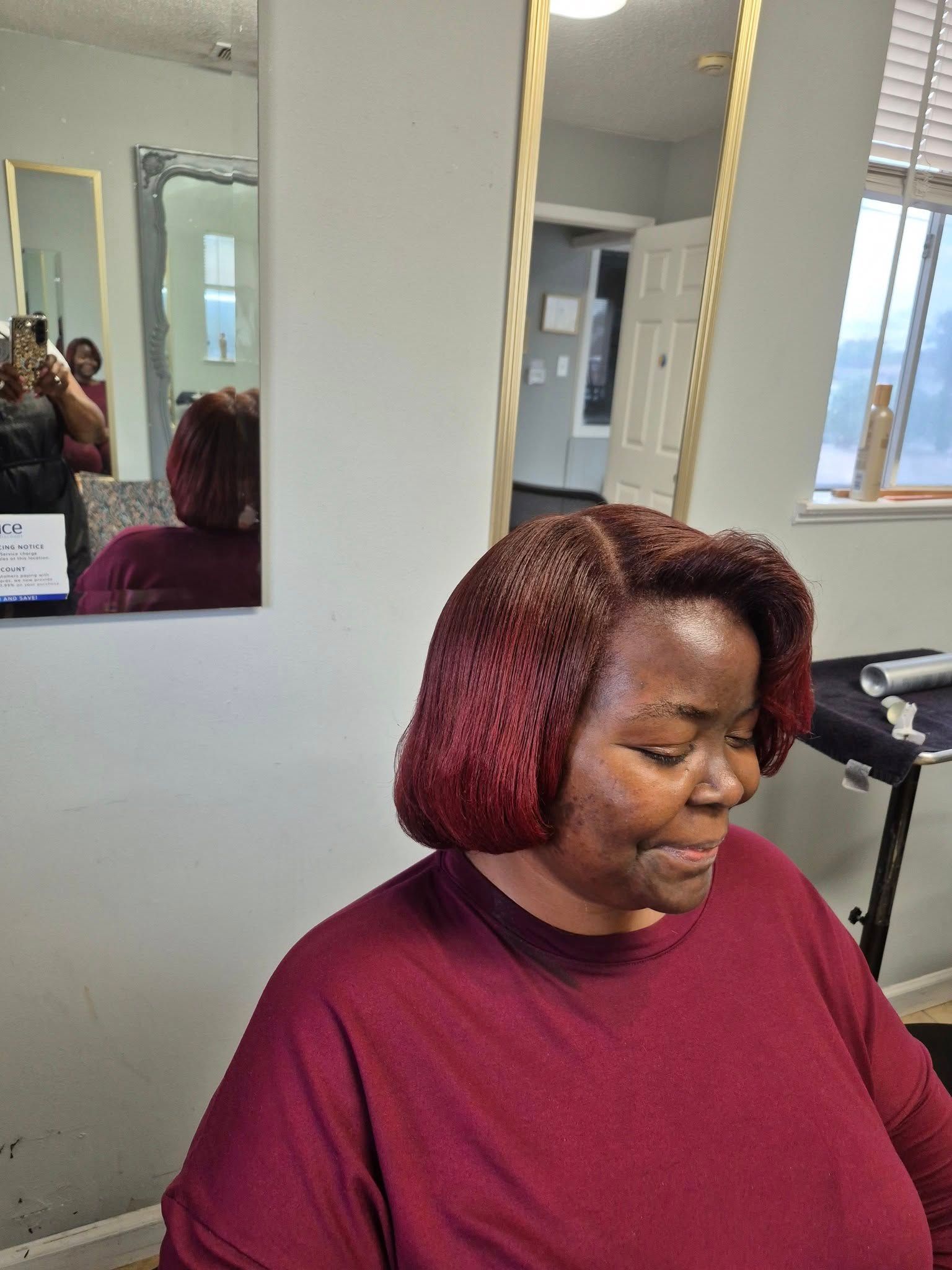 Woman with short, burgundy hair in a salon, wearing a maroon shirt. Mirrors and a window are in the background.