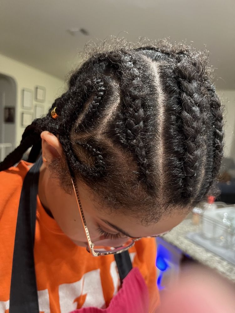 Close-up of a person's head with braided hair styled in cornrows. Orange shirt visible, indoor setting.