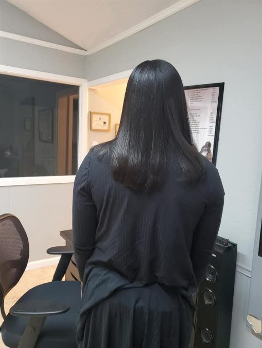 Person with long, dark, straight hair, wearing a black top, standing in a room with gray walls.