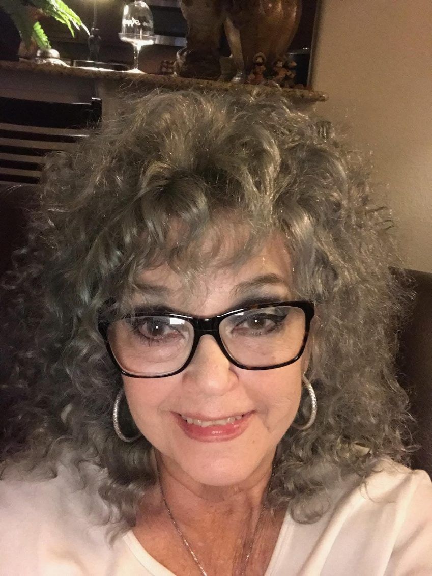 Woman with curly gray hair and glasses smiles at the camera. She wears a white top and hoop earrings.