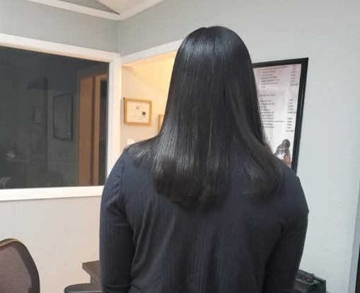 Woman with dark hair in a salon chair, facing away. Backlit by ring light and window.