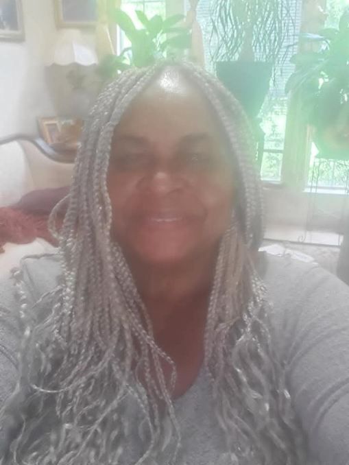 Woman with long silver braids smiles; indoors, soft lighting, gray top, plants in background.