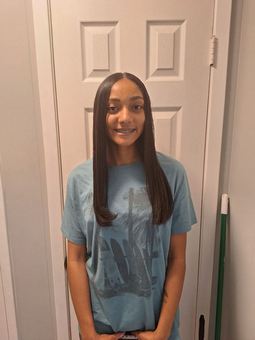 Person with long, dark hair wearing a blue t-shirt stands in front of a white door and smiles.