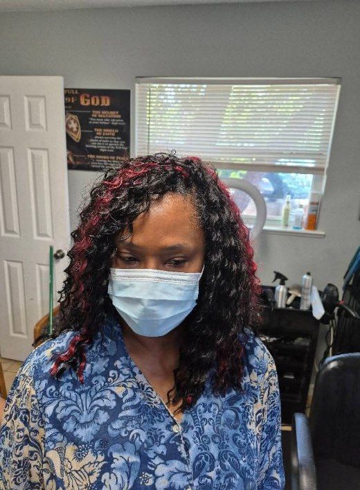 Woman wearing a mask with curly black hair with red highlights. Blue floral shirt. Inside, in front of a window.