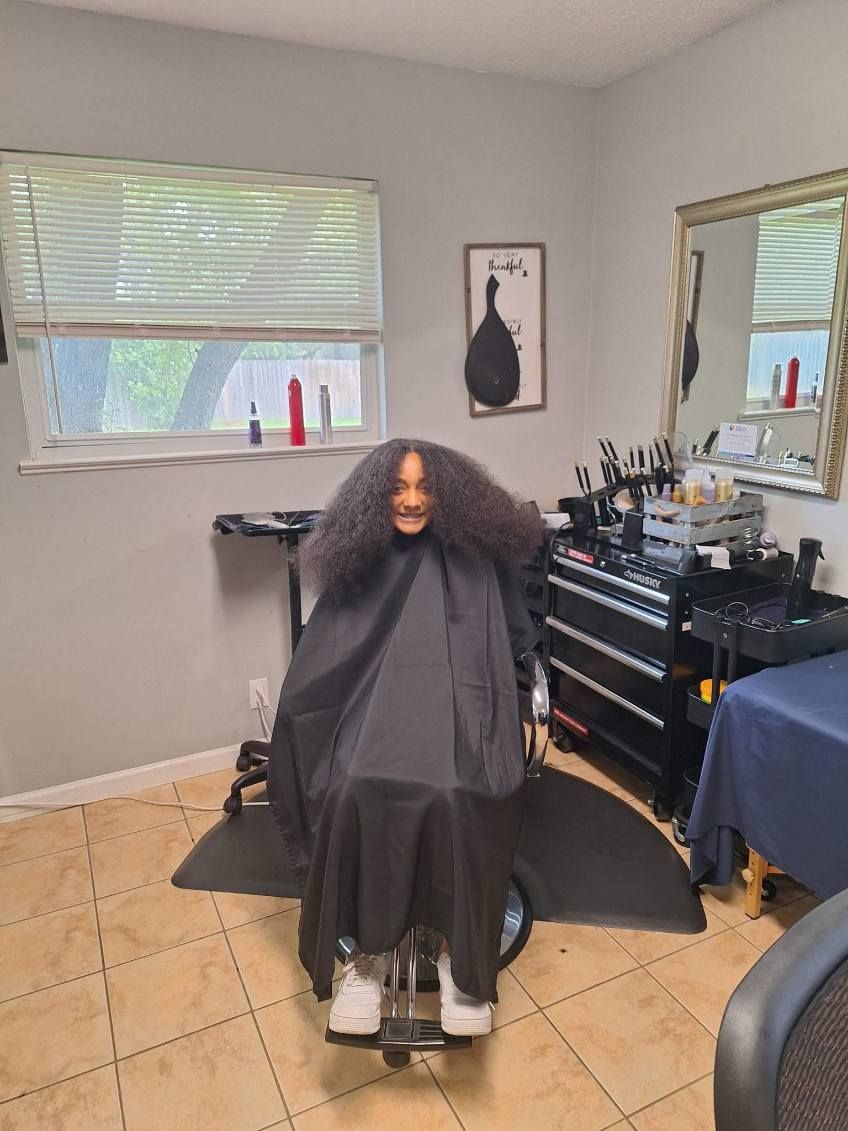 Person in a salon chair with dark hair wearing a black cape. Hairdressing equipment and mirror are in the background.