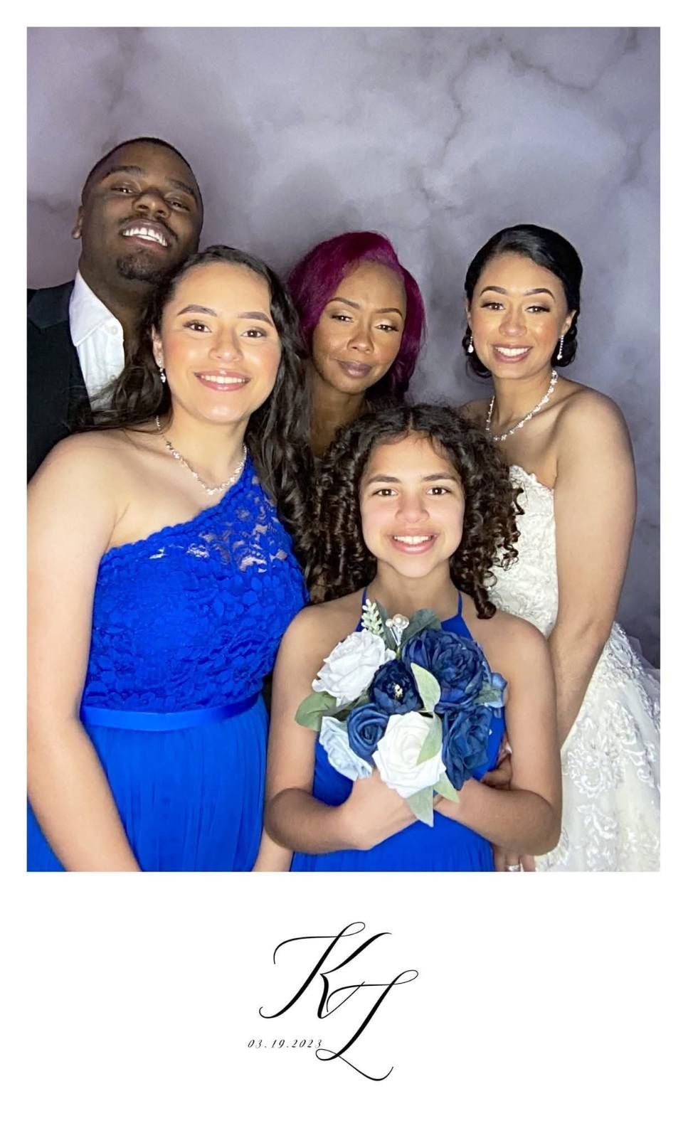 Group photo at event, people smiling, one wearing a bridal gown, others in blue, neutral background.