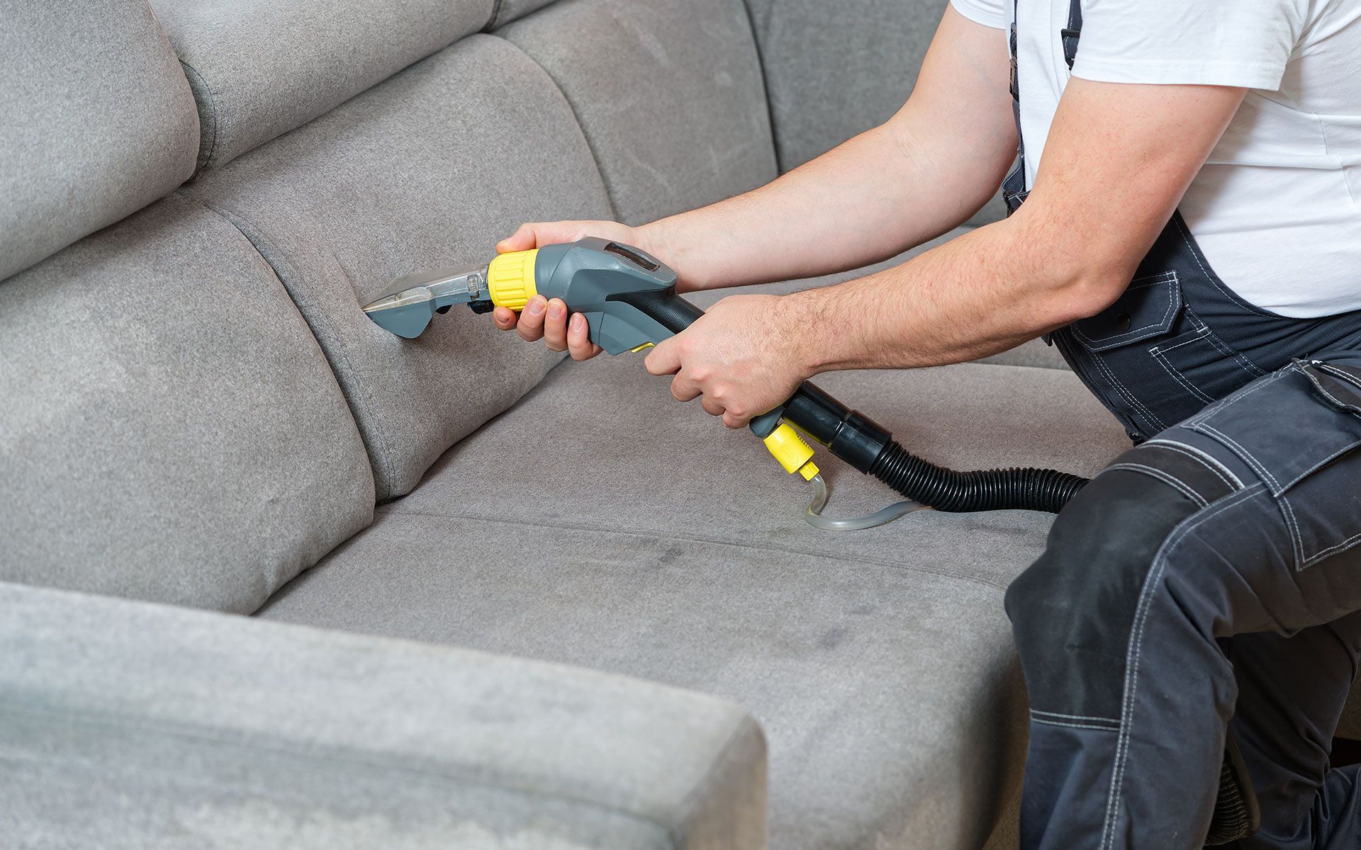Person cleaning a gray sofa with a handheld upholstery cleaner in an indoor setting.