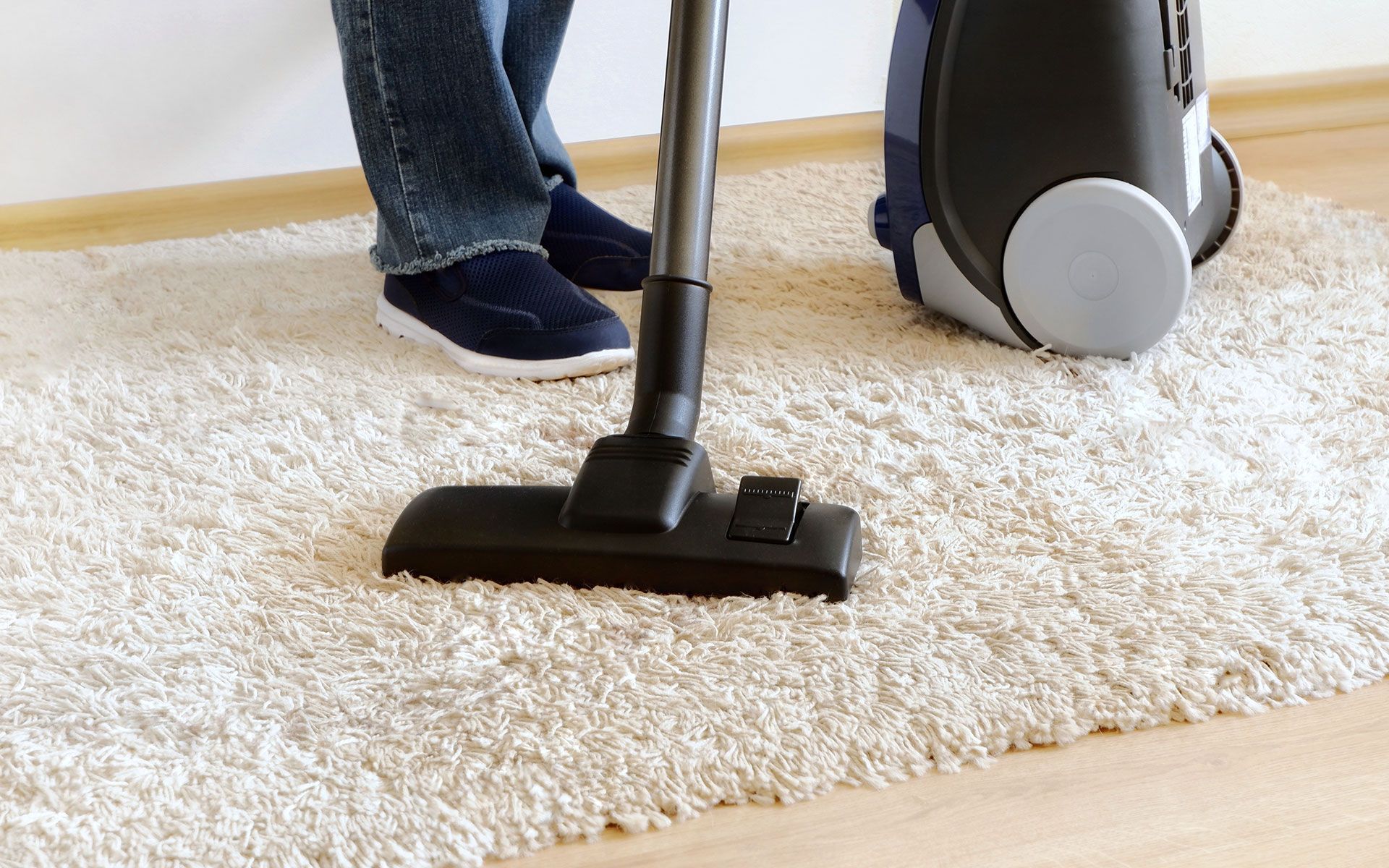 Person vacuuming a cream-colored shag rug with a black vacuum cleaner on a wood floor.