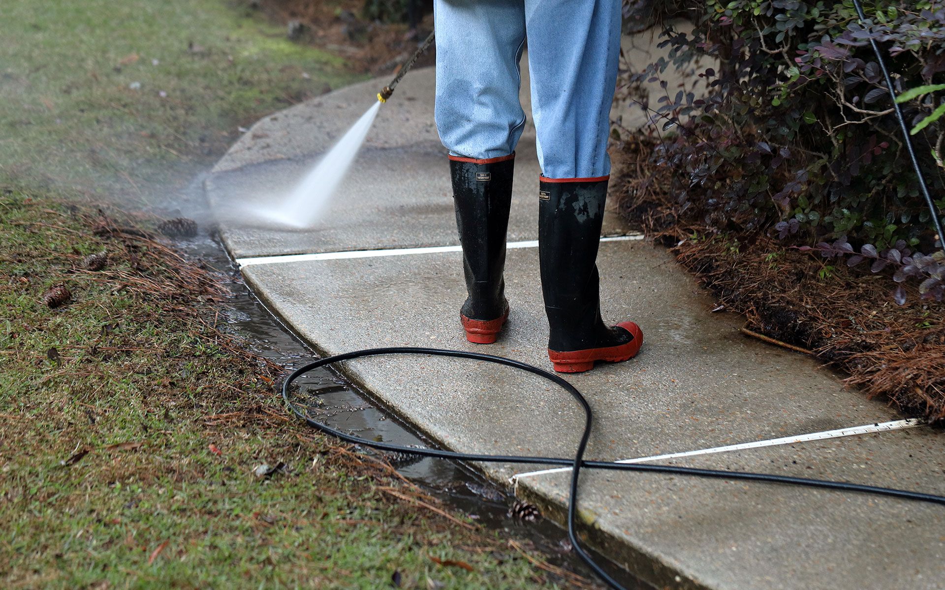 Person pressure washing a concrete sidewalk with water spraying out; grass and bushes on either side.