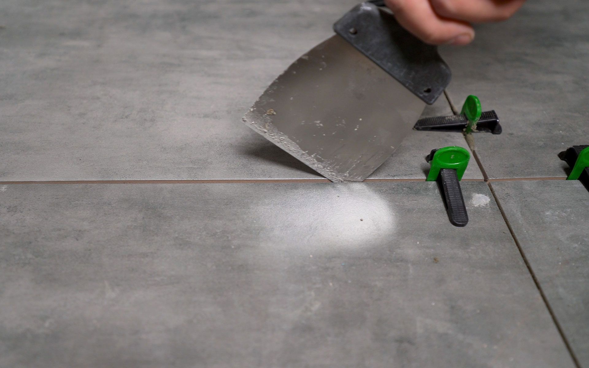 Person spreading grout between gray floor tiles with a metal trowel. Green leveling clips are visible.