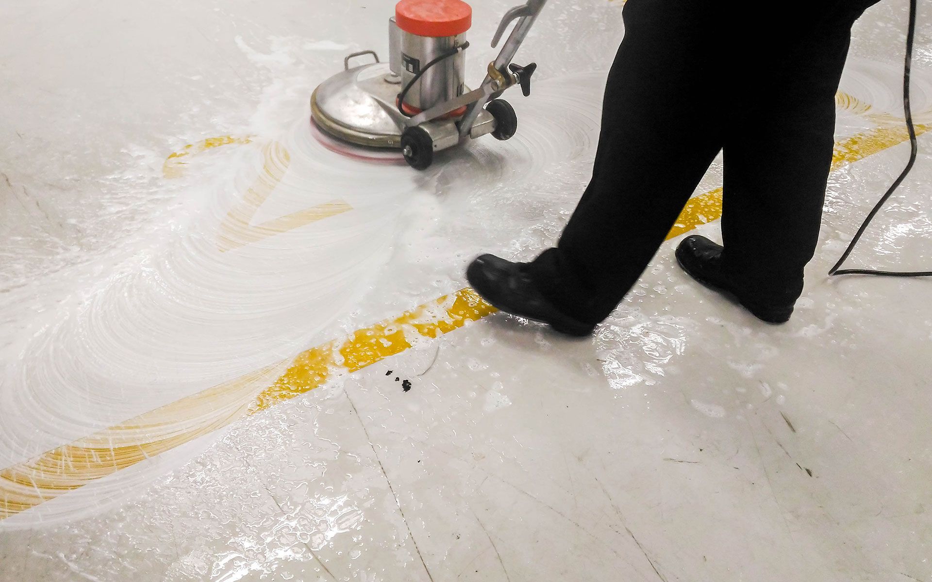 Person using a floor buffer, cleaning a concrete floor with yellow painted lines.