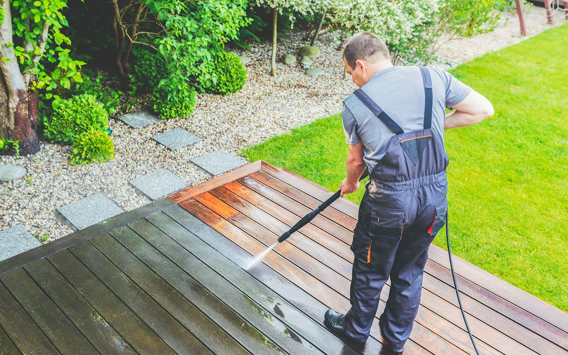 Man power washing a wooden deck, green lawn, and foliage in the background.