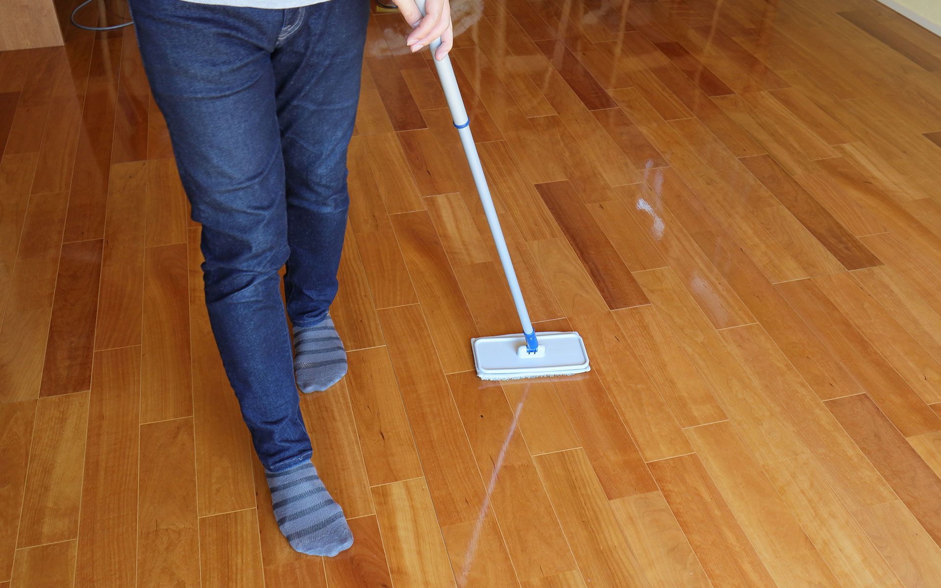Person in jeans mopping a shiny, wooden floor with a white and blue mop.
