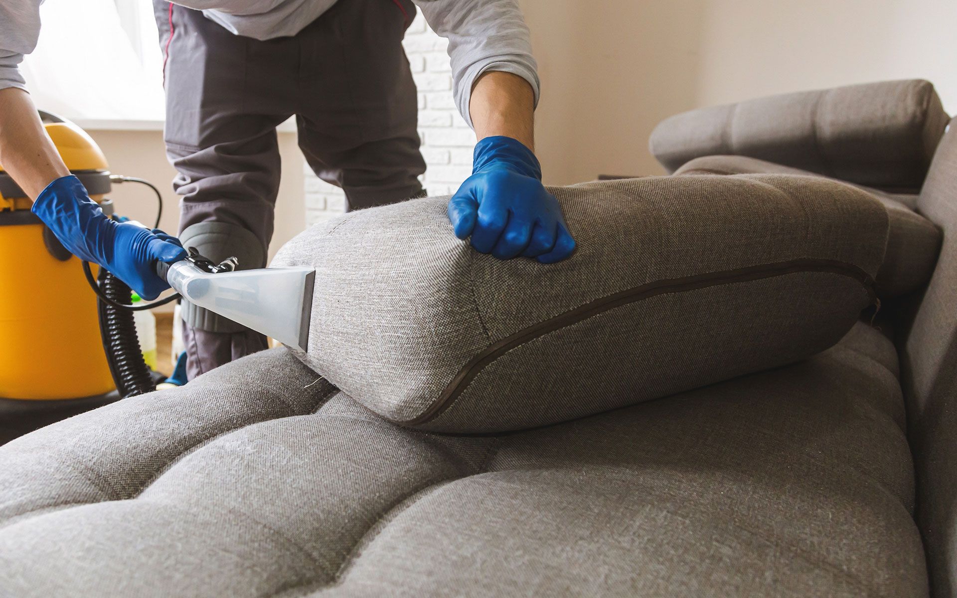 Person in gloves vacuuming a couch pillow with a vacuum cleaner.