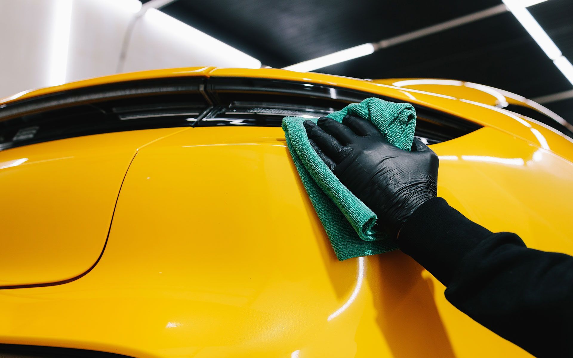 Gloved hand wiping a bright yellow car with a green microfiber cloth.