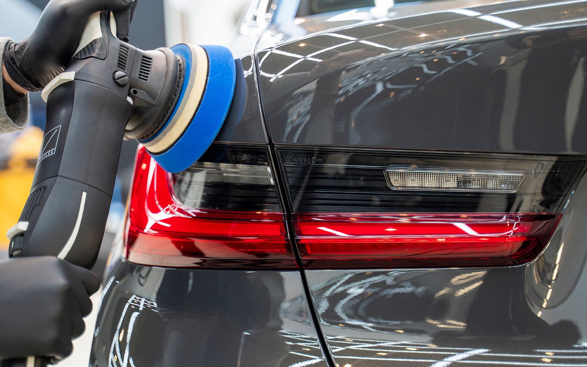 Person polishing a dark gray car with a blue and white buffer near the taillight.