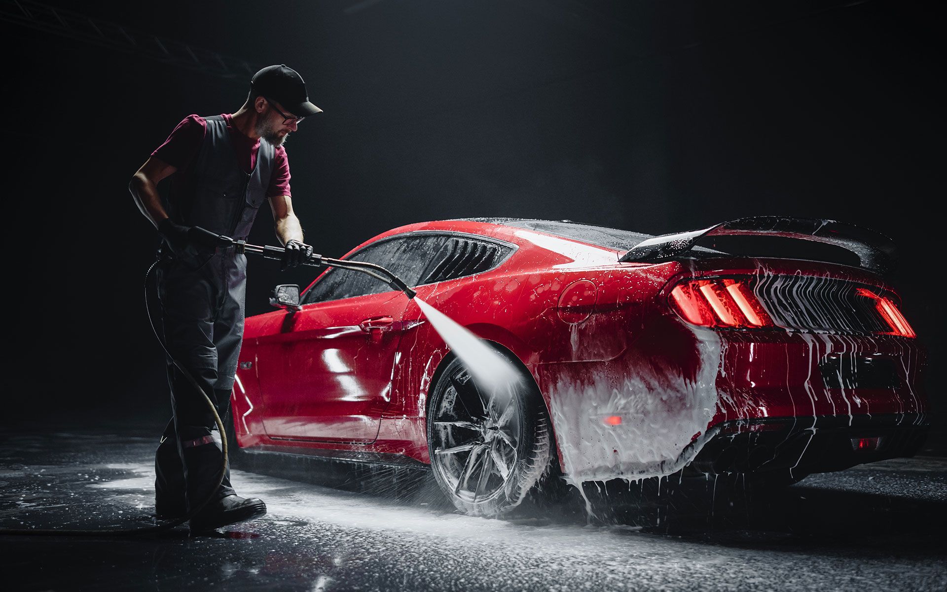 Man washing a red sports car with a pressure washer against a dark backdrop.